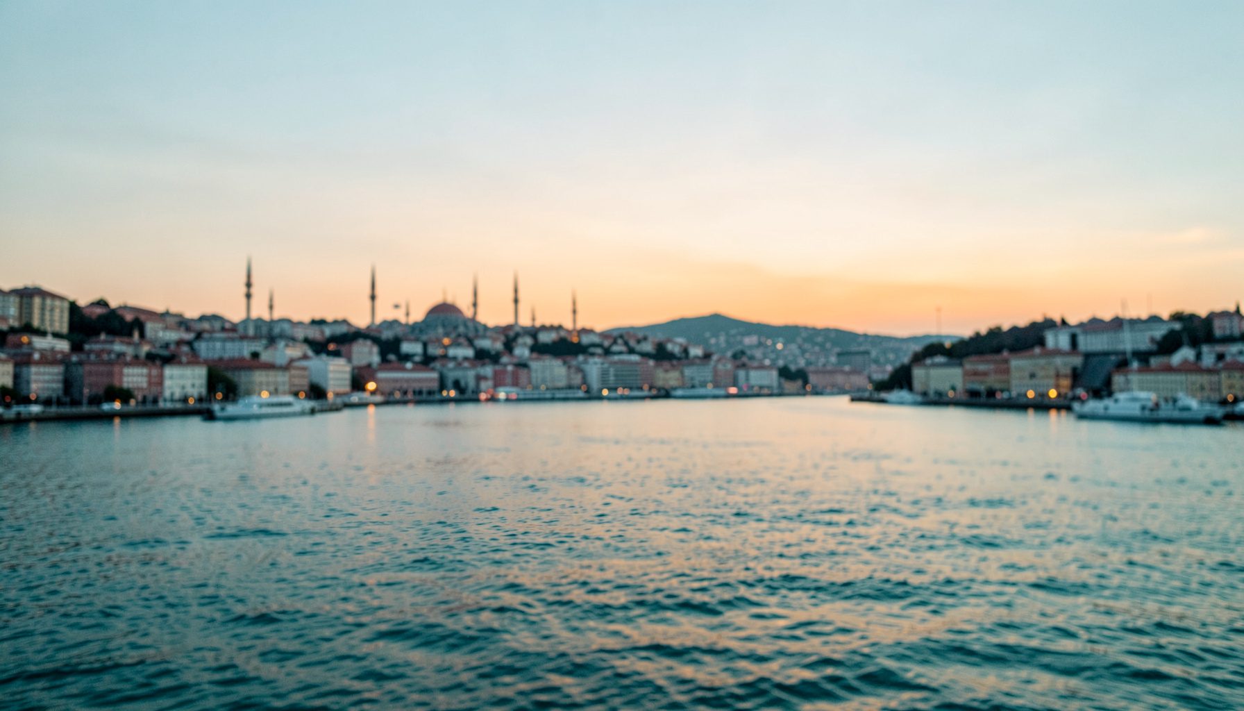 A wide atmospheric photograph of Istanbul's skyline at dusk, with the Bosphorus waterway reflecting soft amber and pale blue light, conveying openness and a sense of civic dialogue