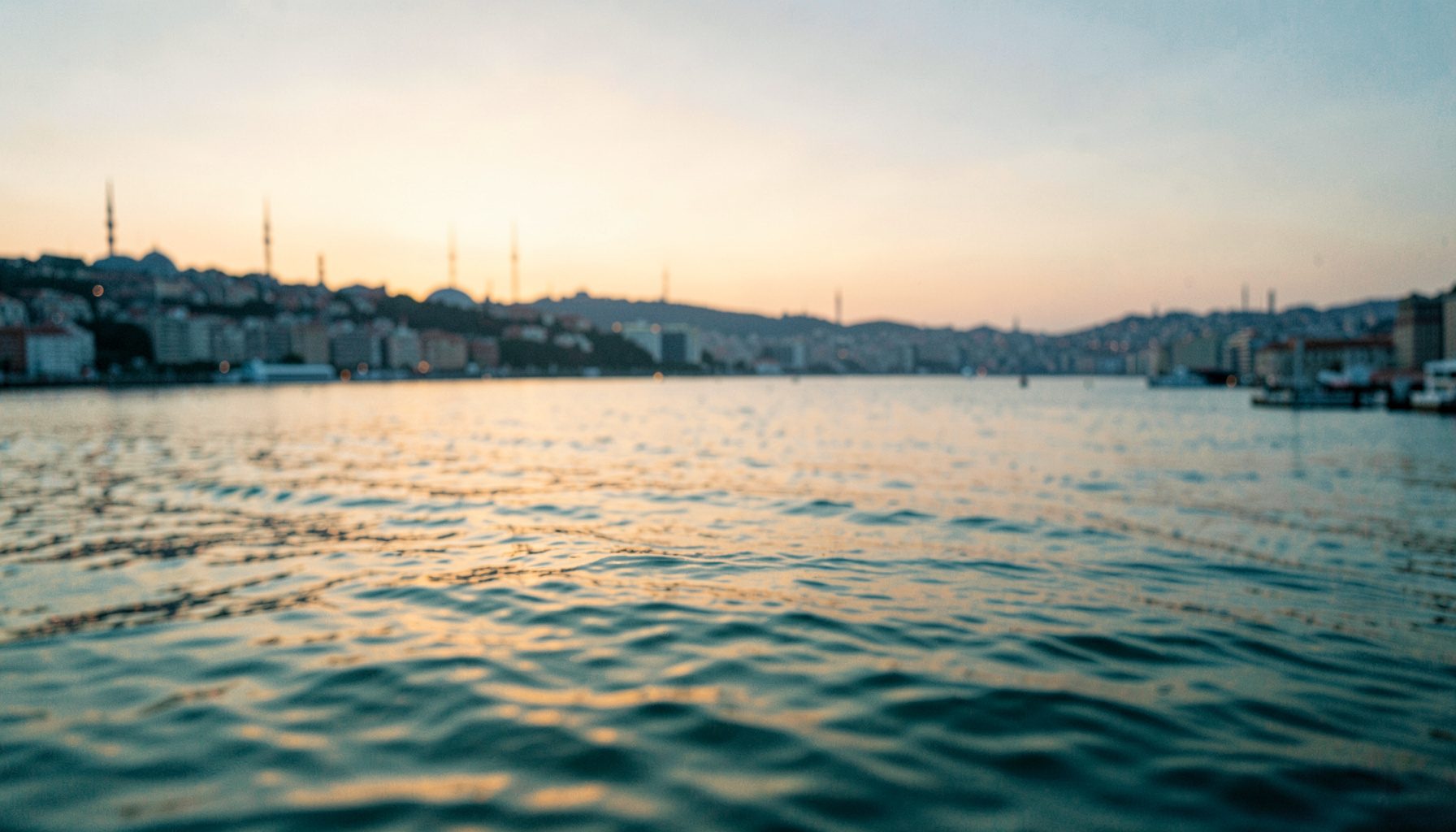 A wide-angle view of Istanbul's Bosphorus shoreline at dusk, with soft golden light reflecting off calm water and the city skyline silhouetted in muted blues and warm amber tones.