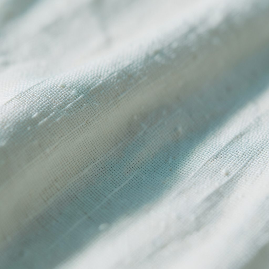 Extreme close-up of smooth cream-colored linen with visible slub texture and gentle crease lines, lit from the side to emphasize the natural fiber grain.