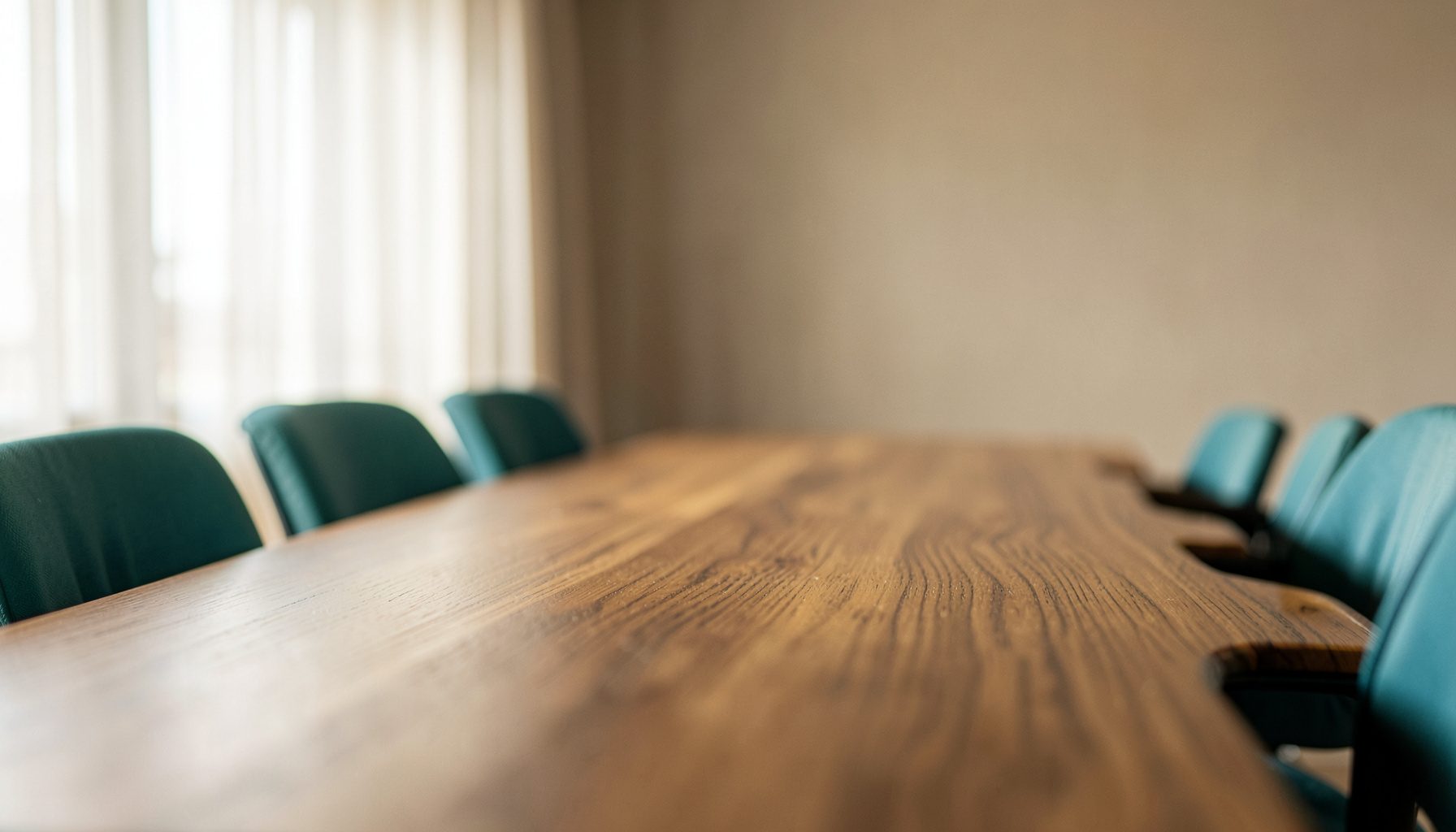 Warmly lit office interior with a long wooden conference table, neutral beige walls, and soft natural daylight filtering through sheer curtains, conveying a calm professional atmosphere.