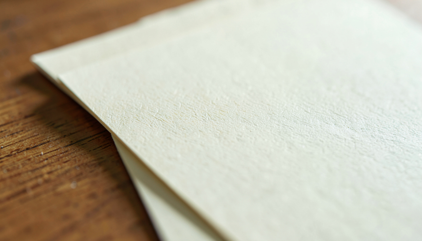 A close-up photograph of smooth cream-colored letter paper with subtle laid texture, resting on a warm wooden desk surface in soft natural daylight.